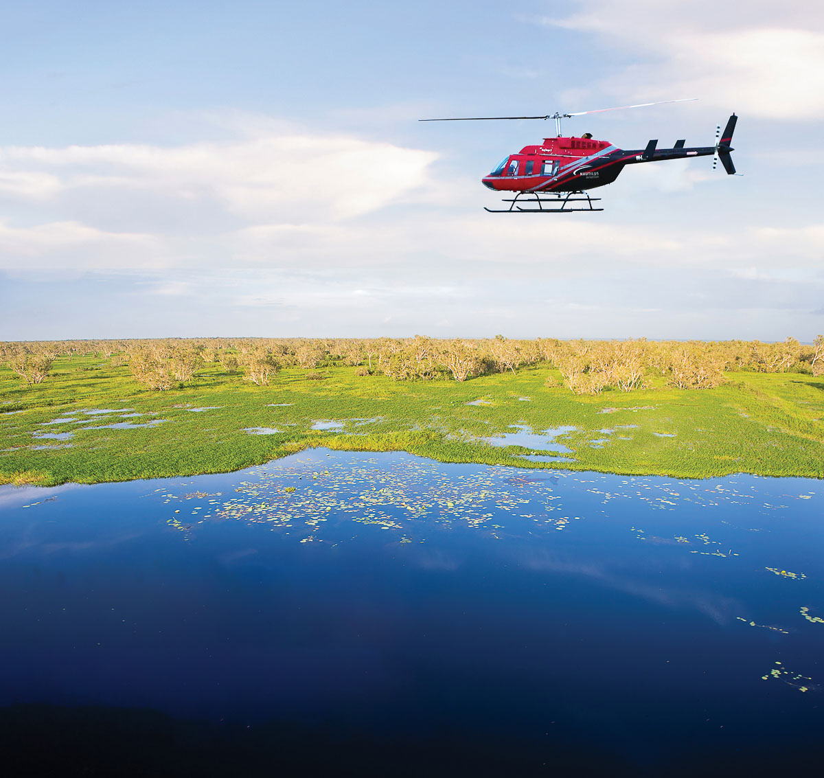 A red helicopter hovers over a lake in the Northern Territory. Darwin Distilling Company's heli-foraging tour.