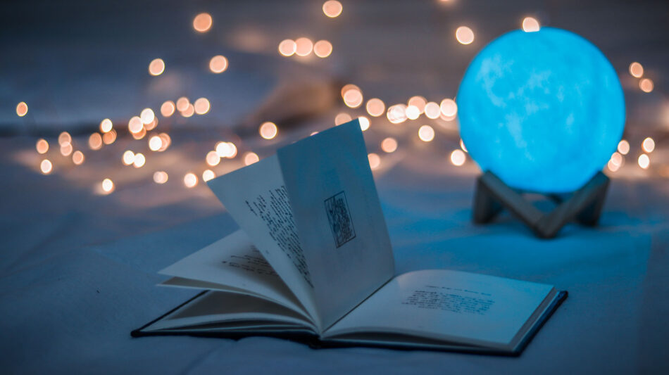 An open book lies on the table at dusk, surrounded by fairy lights and with a glowing moon light in the background.