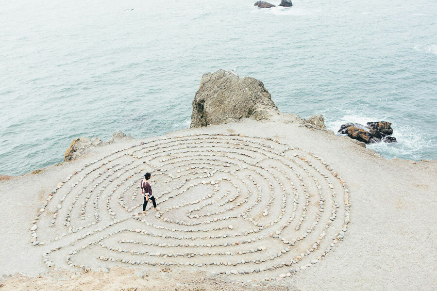 A person approaches a stone labyrinth at the ocean's edge.