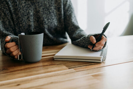 A woman sits at a wooden desk drinking coffee and writing in a journal.