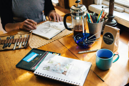 A creative's table loaded with coffee plunger, pens, paper, and other tools.